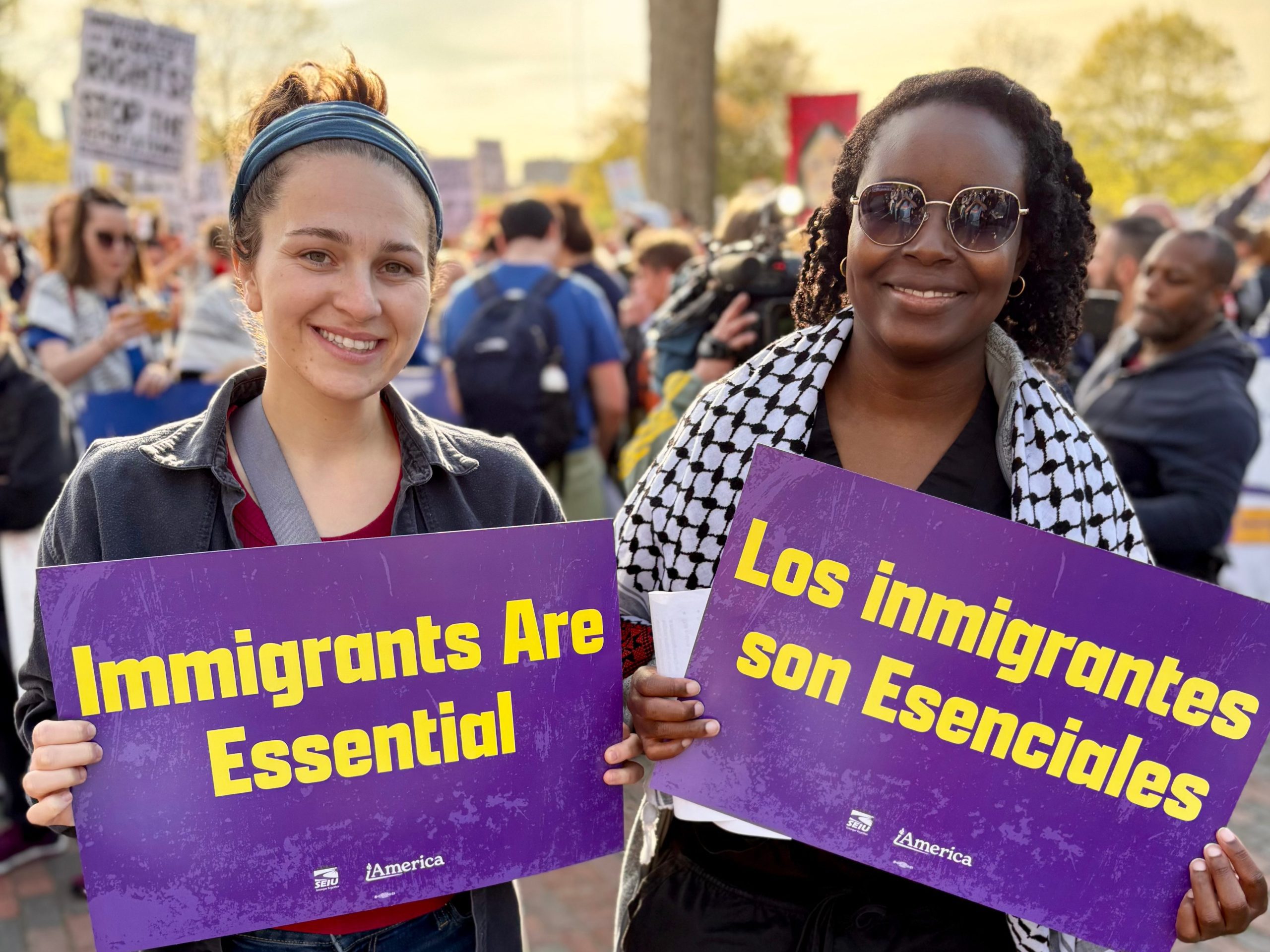 Two women smiling at an outdoor rally, holding matching purple signs with yellow text reading 'Immigrants Are Essential' and 'Los inmigrantes son Esenciales,' with SEIU and ¡America logos at the bottom. A crowd with additional signs is visible in the background.