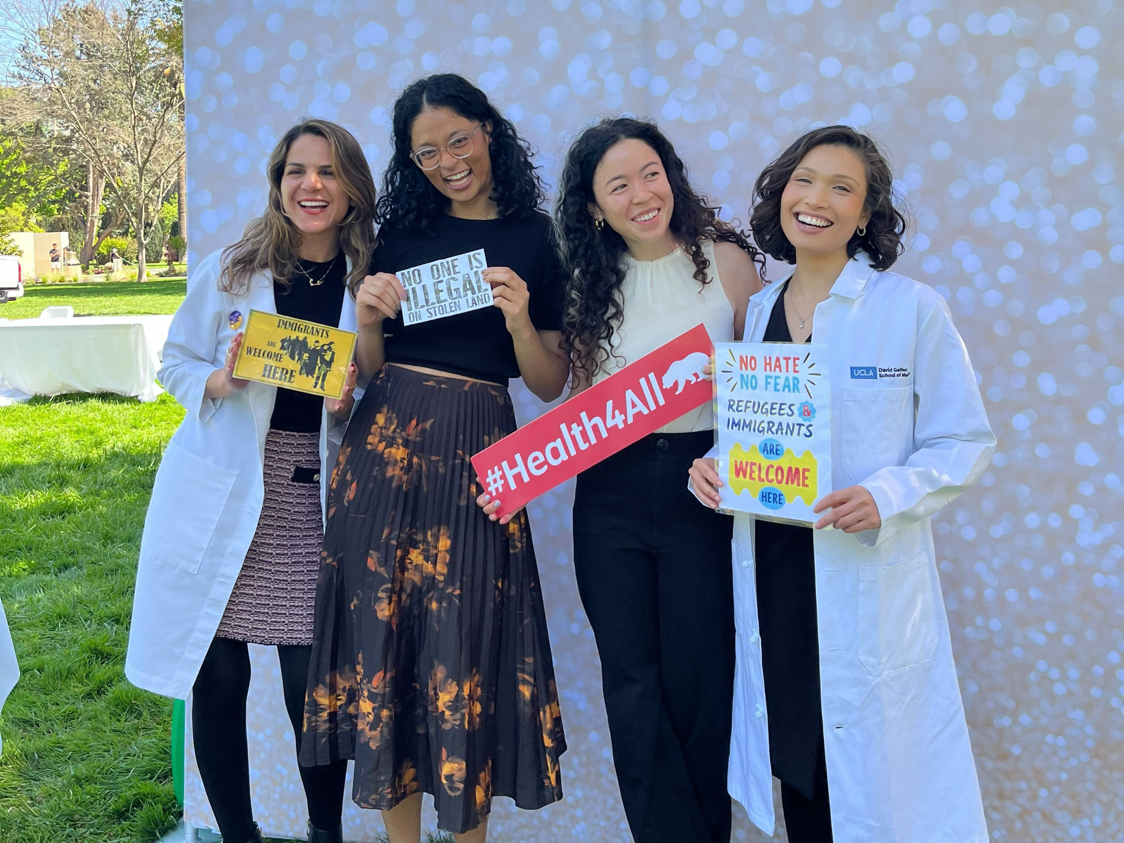 Four smiling women pose outdoors, three in white medical coats, holding signs reading 'Immigrants Are Welcome Here,' 'No One Is Illegal on Stolen Land,' '#Health4All,' and 'No Hate No Fear Refugees & Immigrants Are Welcome Here.'
