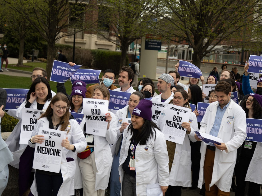 A crowd of people in white medical coats, some wearing masks and purple beanies, hold matching signs reading 'Union-Busting Is Bad Medicine' outdoors near trees and a building.